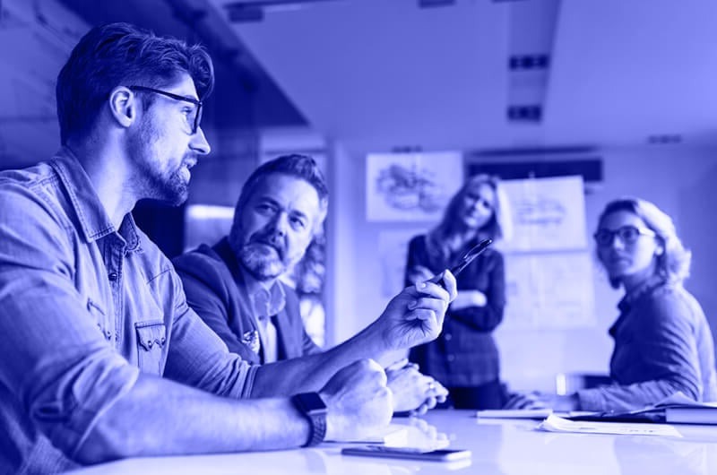 Blue Image of a Man Talking to Three Other People in a Meeting sat at a Table